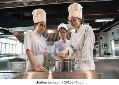 Cooking Class shows a chef instructor stirring a pot as two culinary students observe the demonstration around a stainless steel workstation in a spacious kitchen.