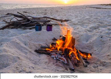 Cooking coffee while traveling on the wild seashore - two mugs stand next to bonfire with small kettle, surrounded by a flame, on the background of setting sun over the sea.
