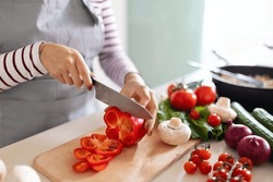 Cooking Concept. Unrecognizable Woman In Grey Apron Making Healthy Dinner, Cropped Of Lady Preparing Delicious Meal At Home, Cutting Vegetables On Chopping Board, Kitchen Interior, Closeup