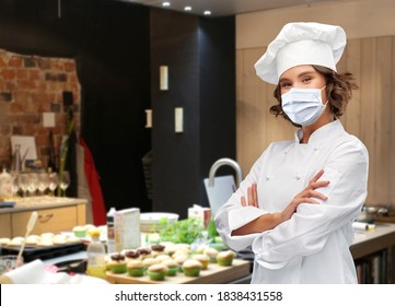 Cooking, Culinary And Health Concept - Female Chef In Toque Wearing Face Protective Medical Mask For Protection From Virus Disease With Crossed Arms Over Restaurant Kitchen Background