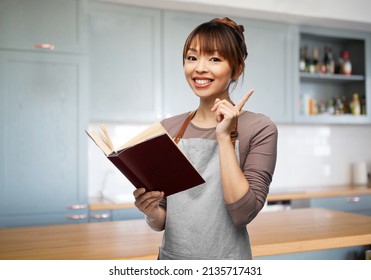 Cooking, Culinary And People Concept - Happy Smiling Woman In Apron With Open Cook Book Over Kitchen Background