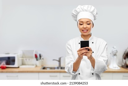 cooking, culinary and people concept - happy smiling female chef in white jacket with smartphone over kitchen background