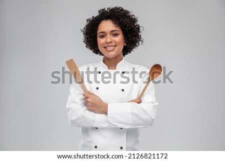 cooking, culinary and people concept - happy smiling female chef in white jacket with wooden spoon and spatula over grey background