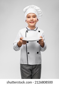 cooking, culinary and profession concept - happy smiling little boy in chef's toque and jacket holding empty plate over grey background