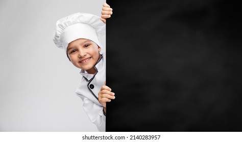 cooking, culinary and profession concept - happy smiling little girl in chef's toque and jacket with black chalkboard over grey background