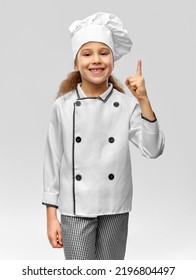 Cooking, Culinary And Profession Concept - Happy Smiling Little Girl In Chef's Toque And Jacket Pointing Finger Up Over Grey Background