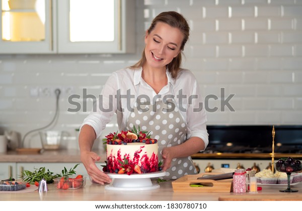 cooking and\
decoration of cake with cream. Young woman pastry chef in the\
kitchen decorating red velvet cake with flowers and berries. Master\
pastry chef completes work on a birthday\
cake