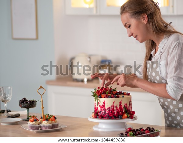 cooking and\
decoration of cake with cream. Young woman pastry chef in the\
kitchen decorating red velvet cake with flowers and berries. Master\
pastry chef completes work on a birthday\
cake