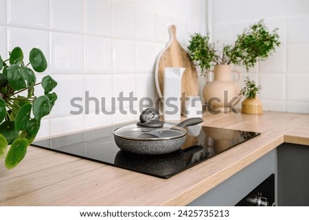 Cooking dinner concept. Household appliance. Detail in kitchen interior, induction glass ceramic stove on wooden countertop, frying pan with lid, plant in vase, cutting board and white tile on wall.