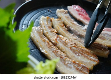 Cooking Grilled Pork Belly Or Samgyeopsal (Korean Name) In Korean BBQ Non-Stick Grill Pan With Kitchen Tongs, Blurred Foreground Of Fresh Green Vegetables. People Stay Home And Enjoy Cooking Lifestyle