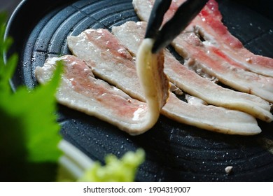 Cooking Grilled Pork Belly Or Samgyeopsal (Korean Name) In Korean BBQ Non-Stick Grill Pan With Kitchen Tongs, Blurred Foreground Of Fresh Green Vegetables. People Stay Home And Enjoy Cooking Lifestyle