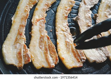 Cooking Grilled Pork Belly Or Samgyeopsal (Korean Name) In Korean BBQ Non-Stick Grill Pan With Kitchen Tongs. People Stay Home And Enjoy Cooking Lifestyle Theme. (close Up Shot, Selective Focus)