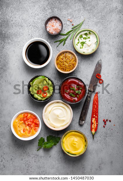 Cooking healthy sauces\
and dressings concept. Different dips in bowls on rustic concrete\
background, ingredients, herbs, pepper chili and kitchen knife, top\
view, close-up