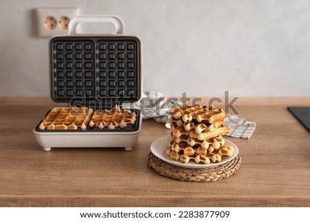 Cooking homemade waffles. A waffle iron with Belgian waffles and a stack of homemade waffles on a wooden kitchen table. 