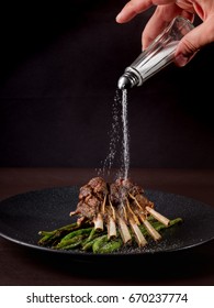 Cooking Lamb Ribs With Grilled Vegetables On Food Dark Background With Chef Cook Hand And Salt. Top View On Stone Black Plate. Vertical Photo.