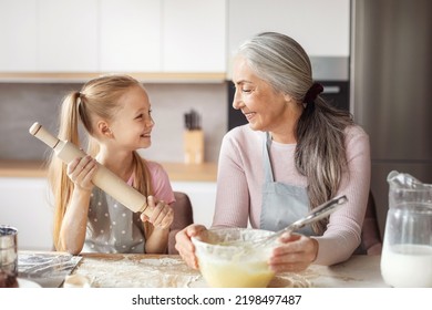 Cooking Lesson. Smiling European Little Granddaughter And Old Grandma In Aprons Make Dough With Rolling Pin, Have Fun In Kitchen Interior. Prepare Homemade Food Together At Home, Love And Relationship