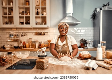 Cooking with love. Plump african american woman expertly molding dough sprinkled with flour on wooden kitchen surface. Smiling homemaker in turban and apron baking handmade bread at modern house. - Powered by Shutterstock