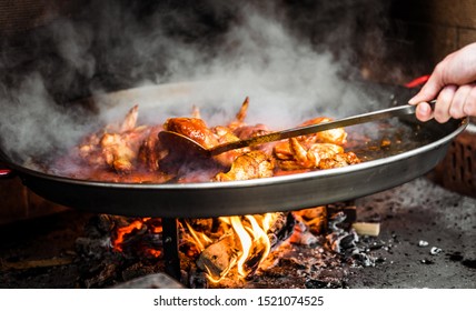 Cooking And Making A Traditional Spanish Paella Over Open Fire. Traditional Way Of Preparing Valencian Paella  With Fire Wood And Flames In A Big Pan. Made From Chicken And Rabbit Meat In Fireplace.