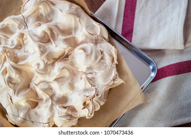 Cooking The Meringue Base For A Pavlova Dessert. Fresh Meringue, With Crisp Golden Top, On Baking Parchment And A Stainless Steel Baking Tray. Overhead View On Wooden Table With Cloth.