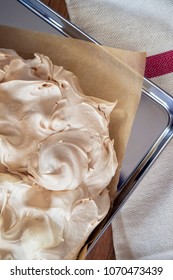 Cooking The Meringue Base For A Pavlova Dessert. Fresh Meringue, With Crisp Golden Top, On Baking Parchment And A Stainless Steel Baking Tray. Overhead View On Wooden Table With Cloth.