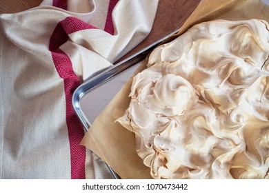 Cooking The Meringue Base For A Pavlova Dessert. Fresh Meringue, With Crisp Golden Top, On Baking Parchment And A Stainless Steel Baking Tray. Overhead View On Wooden Table With Cloth.