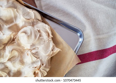 Cooking The Meringue Base For A Pavlova Dessert. Fresh Meringue, With Crisp Golden Top, On Baking Parchment And A Stainless Steel Baking Tray. Overhead View On Wooden Table With Cloth.