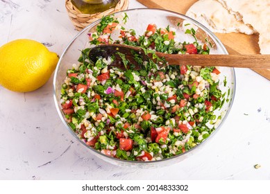 Cooking oriental summer arabic vegetarian bulgur salad Tabouleh. Mixed bulgur, chopped tomato, onion and herbs with a wooden spoon in a glass bowl. Top view with lemon and pita on wooden kitchen board