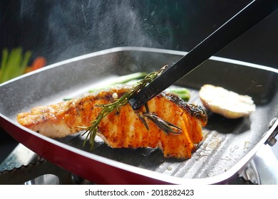 Cooking pan seared salmon moment, sliced salmon with rosemary in nonstick teflon coating pan with garlic, vegetables, black kitchen tongs. Ketogenic diet meal preparation theme. (selective focus)