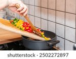 A cooking pot on gas fire. Person putting fresh cut paprikas from a wooden cutting board into a cooking pot on gas stove. The gas fire is on. Healthy food cooking, home made healthy dinner. Fresh vege