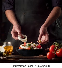 Cooking Process Of Greek Salad For A Restaurant Menu. Food Composition Of A Mediterranean Cuisine In Rustic Style. Chef Put The Onion And Olives In A Black Bowl And Mixed Of All Vegetables In A Salad.