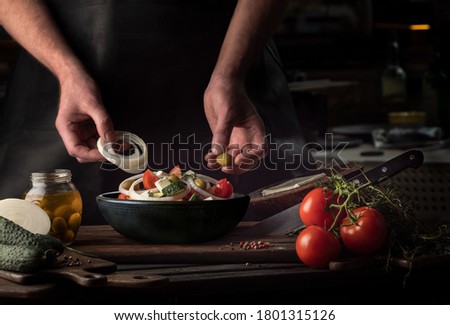 Cooking process of Greek Salad for a restaurant menu. Food composition of a Mediterranean cuisine in rustic style. Chef put the onion and olives in a black bowl and mixed of all vegetables in a salad.