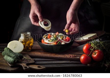 Cooking process of Greek Salad for a restaurant menu. Food composition of a Mediterranean cuisine in rustic style. Chef put the onion and olives in a black bowl and mixed of all vegetables in a salad.