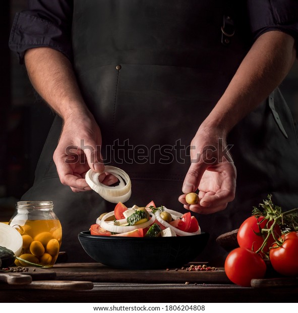 Cooking process of Greek Salad for a restaurant\
menu. Food composition of a Mediterranean cuisine in rustic style.\
Chef put the onion and olives in a black bowl and mixed of all\
vegetables in a salad.