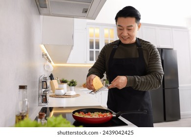 Cooking process. Man grating cheese into frying pan in kitchen