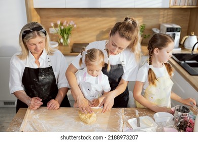 Cooking Process In Modern Light Kitchen With Window. Young Mother And Mother In Law Teaching Little Girl Kneading Dough. Older Sister Turned Away Frome Everyone Examimg Kitchen Tools.