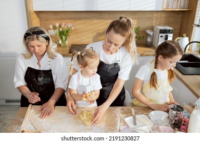 Cooking Process In Modern Light Kitchen With Window. Young Mother And Mother In Law Teaching Little Girl Kneading Dough. Older Sister Turned Away Frome Everyone Examimg Kitchen Tools.