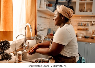 Cooking routine. Side view of oversized woman in headband and apron washing hands in sink before working with food at home kitchen. Smiling african american housewife preparing for domestic cooking.