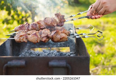 Cooking shish kebab on the grill with smoke, the man flips the skewers brazier. Fresh ruddy meat for barbecue grilled on fire outdoor. Picnic in nature - BBQ. Shashlik is fried in summer sunny garden