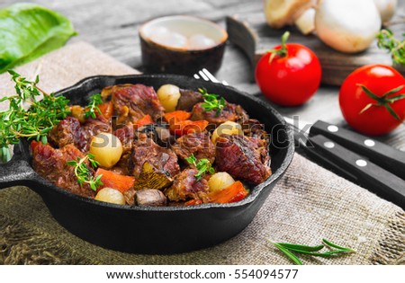 Cooking Stew meat in Burgundy (Beef Bourguignon) in cast iron frying pan with carrots, onions. Spices for Beef Bourguignon thyme, cherry tomatoes, mushrooms champignons. Gray wooden background.