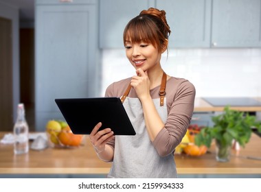 cooking, technology and people concept - happy smiling female chef or waitress in apron with tablet pc computer over home kitchen background