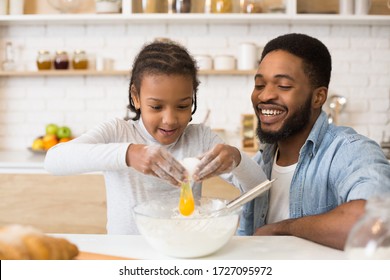 Cooking Together At Home. Excited African Girl Adding Egg To Cookies Dough, Cheerful Dad Watching Her, Copy Space