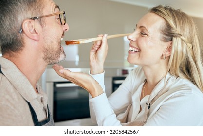 Cooking Woman Feed Man To Taste Food, Meal And Lunch In Kitchen At Home. Happy, Smile And Care Wife Giving, Hungry And Tasting Husband Mouth Spoon Of Tomato Sauce While Couple Eating Dinner With Love