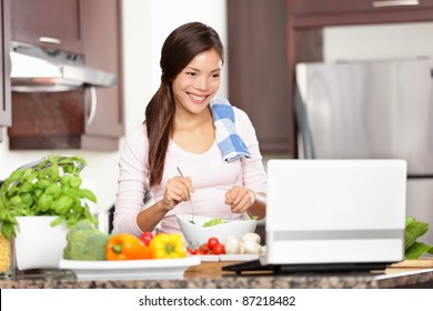 Cooking Woman Looking At Computer While Preparing Food In Kitchen. Beautiful Young Multiracial Woman Reading Cooking Recipe Or Watching Show While Making Salad.