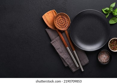 Cooking wooden utensils, black empty plate, basil leaves and spices on dark stone background. Abstract food background. Top view of dark rustic kitchen table with wooden cooking spoon, frame. Mockup.
