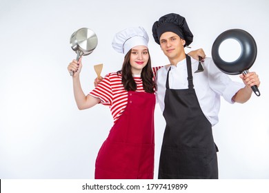 Cooks. A Man And A Woman In Cooks Caps. Two Students Of Chef Cook Show Pans. Concept - Training In Cooking. Two Chefs Stand In An Embrace On A White Background. Students Study To Become A Chef Cook