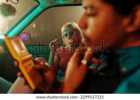 A cool 90s fashioned couple is in the car spending time together. The man is playing a vintage video game and the woman is blowing bubbles with a chewing gum and looking at the camera.