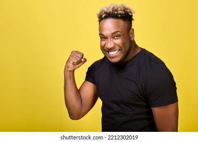 Cool African American in a stylish T-shirt is very happy and satisfied, making a winner's gesture with his hand raised, clenched into a fist. Dark-skinned strong man smiles and screams for success - Powered by Shutterstock