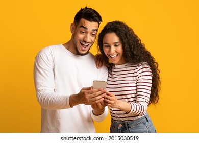 Cool App. Excited arab couple looking at smartphone screen in hands, joyful middle eastern spouses browsing new application while standing together over yellow studio background, copy space