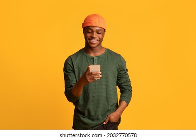 Cool App. Happy Young Black Guy Using Smartphone, Browsing New Application Or Messaging With Friends Online On Mobile Phone, Cheerful African Man Standing Over Yellow Studio Background, Copy Space