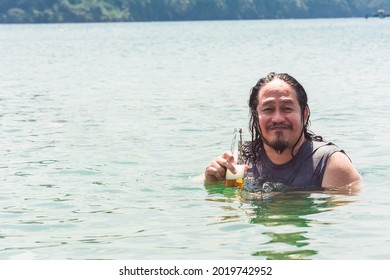 A Cool Asian Guy With Long Hair And Facial Hair Enjoys A Cold Bottle Of Beer While Chilling In Chest Deep Waters. Unwinding At The Beach.